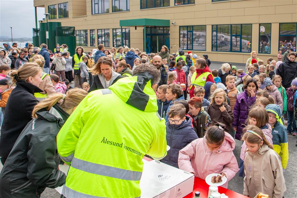 Voksen med anleggsjakke med Ålesund kommune-logo serverer kake til barn - Klikk for stort bilete
