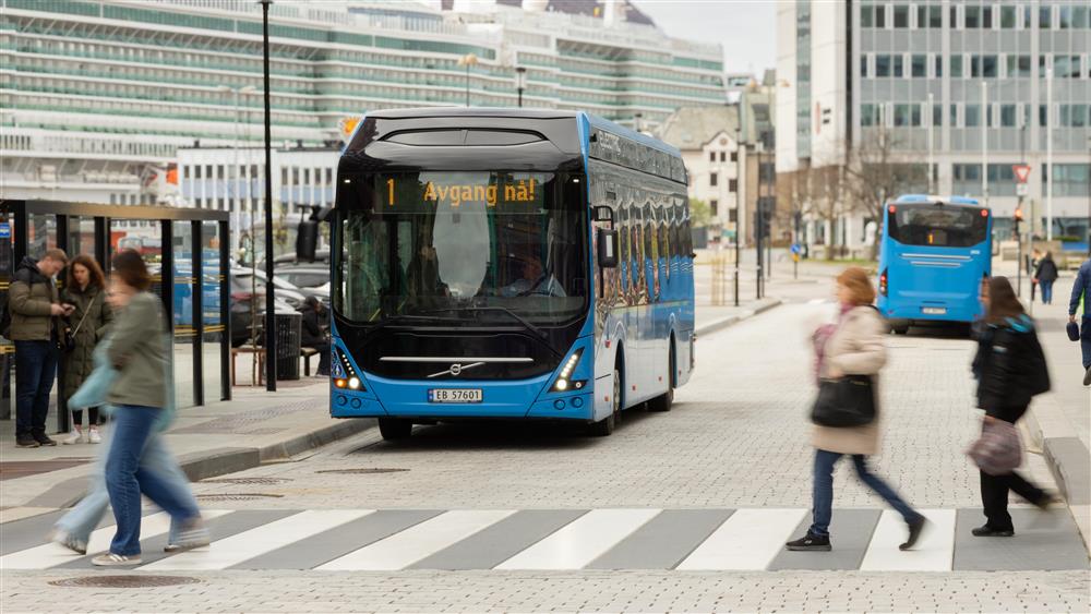 Mennesker som går på gangfelt, og busser i begge retninger på kollektivknutepunktet.  - Klikk for stort bilete